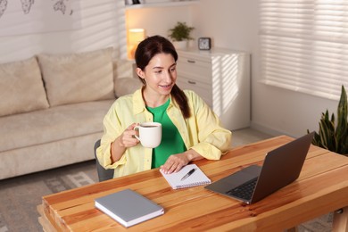 Photo of Young woman with cup of coffee working on laptop at desk in home office