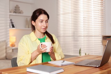 Photo of Young woman with cup of coffee working on laptop at desk in home office. Space for text