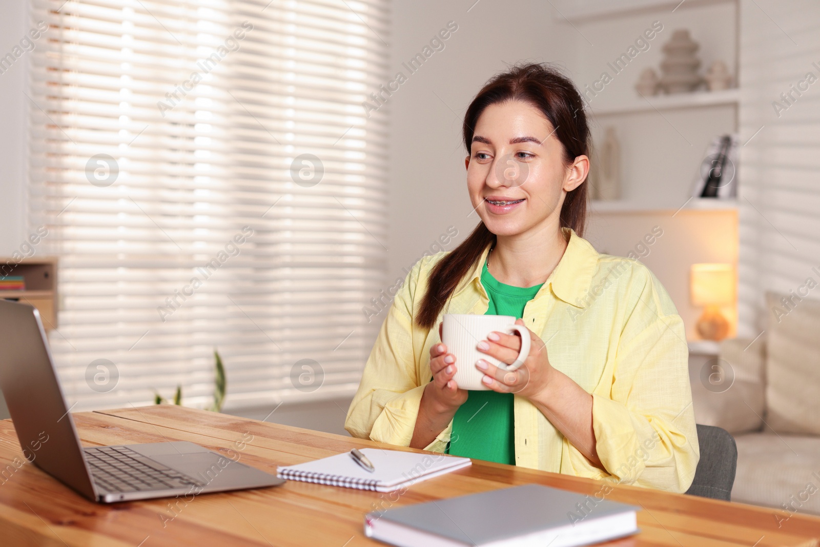 Photo of Young woman with cup of coffee working on laptop at desk in home office. Space for text