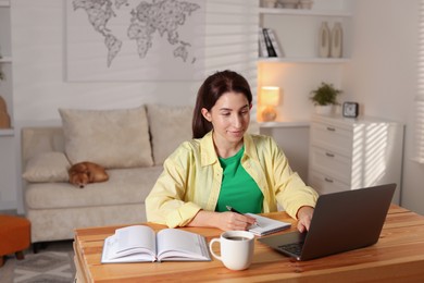 Photo of Young woman working with laptop at desk in home office while her cute dog sleeping on sofa, selective focus