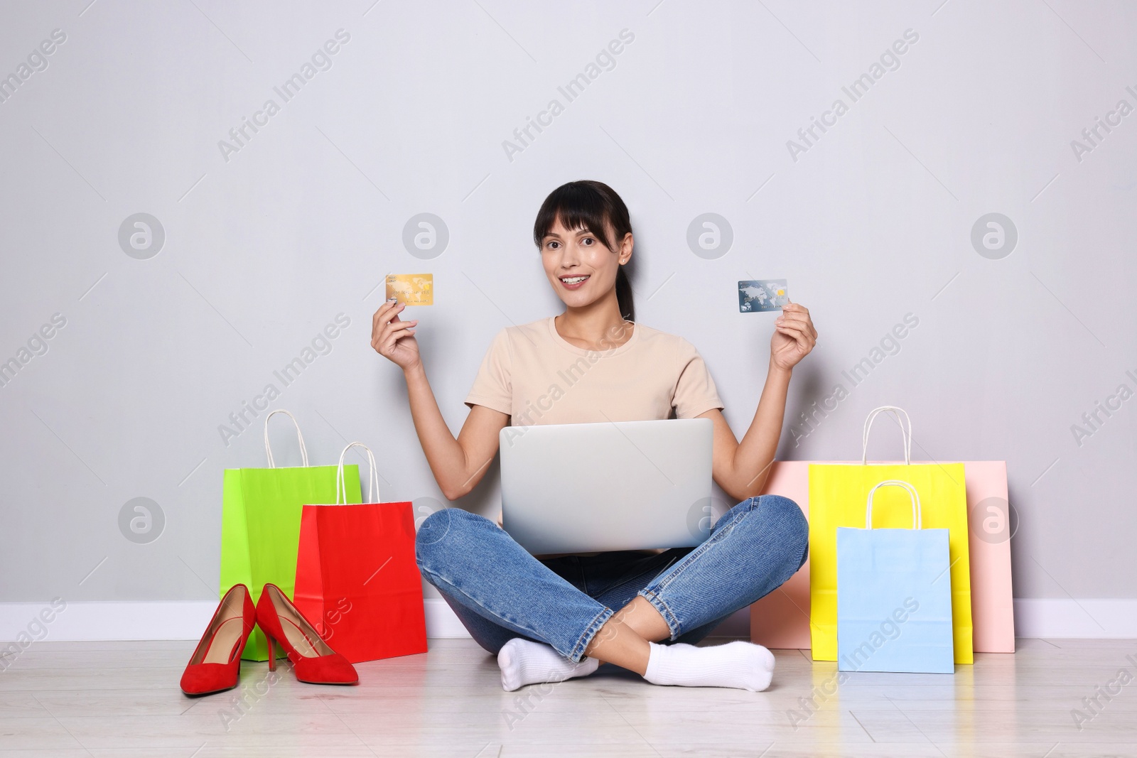 Photo of Internet shopping. Happy woman with credit cards, laptop and colorful bags sitting near grey wall