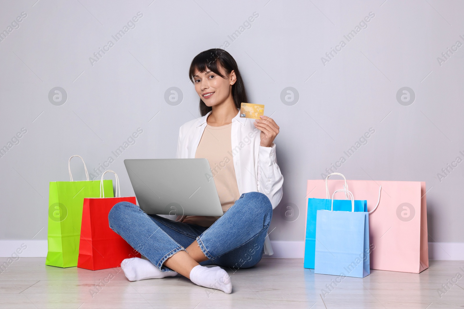 Photo of Internet shopping. Happy woman with credit card, laptop and colorful bags sitting near grey wall