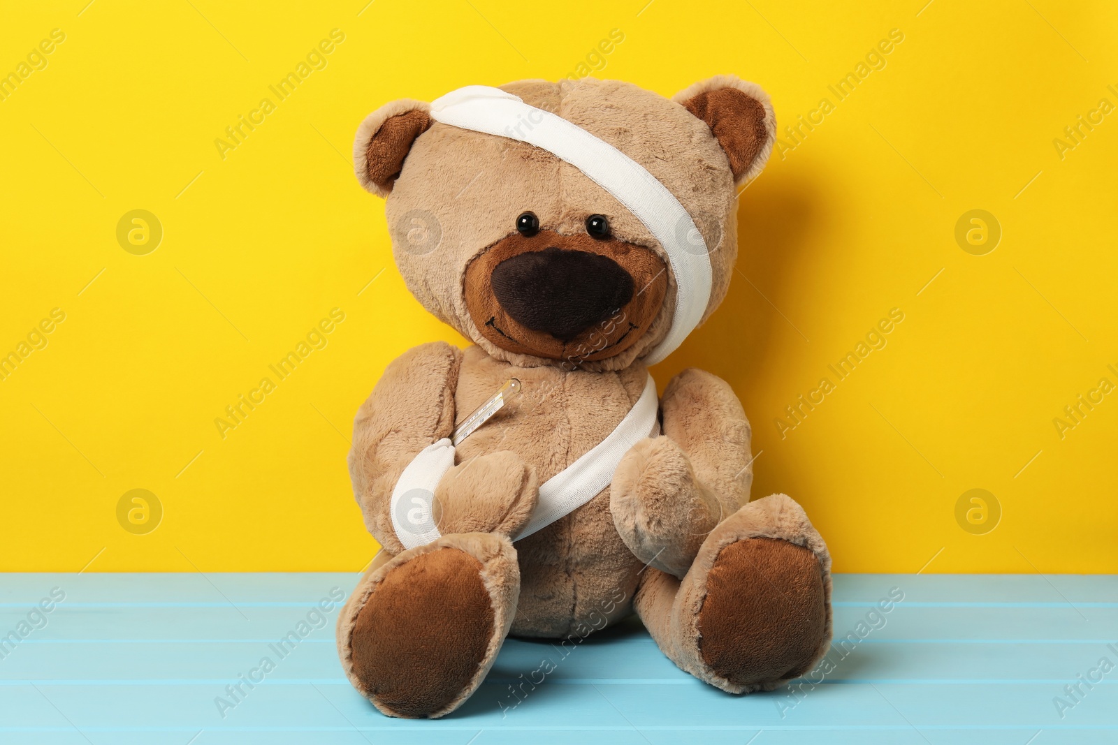 Teddy bear with bandages and thermometer on light blue wooden table against yellow background Photo of Teddy bear with bandages and thermometer on light blue wooden table against yellow background