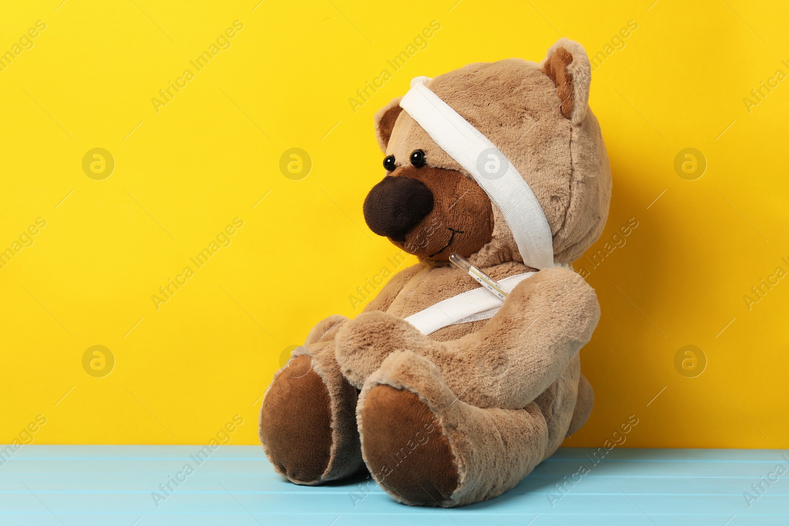 Teddy bear with bandages and thermometer on light blue wooden table against yellow background Photo of Teddy bear with bandages and thermometer on light blue wooden table against yellow background