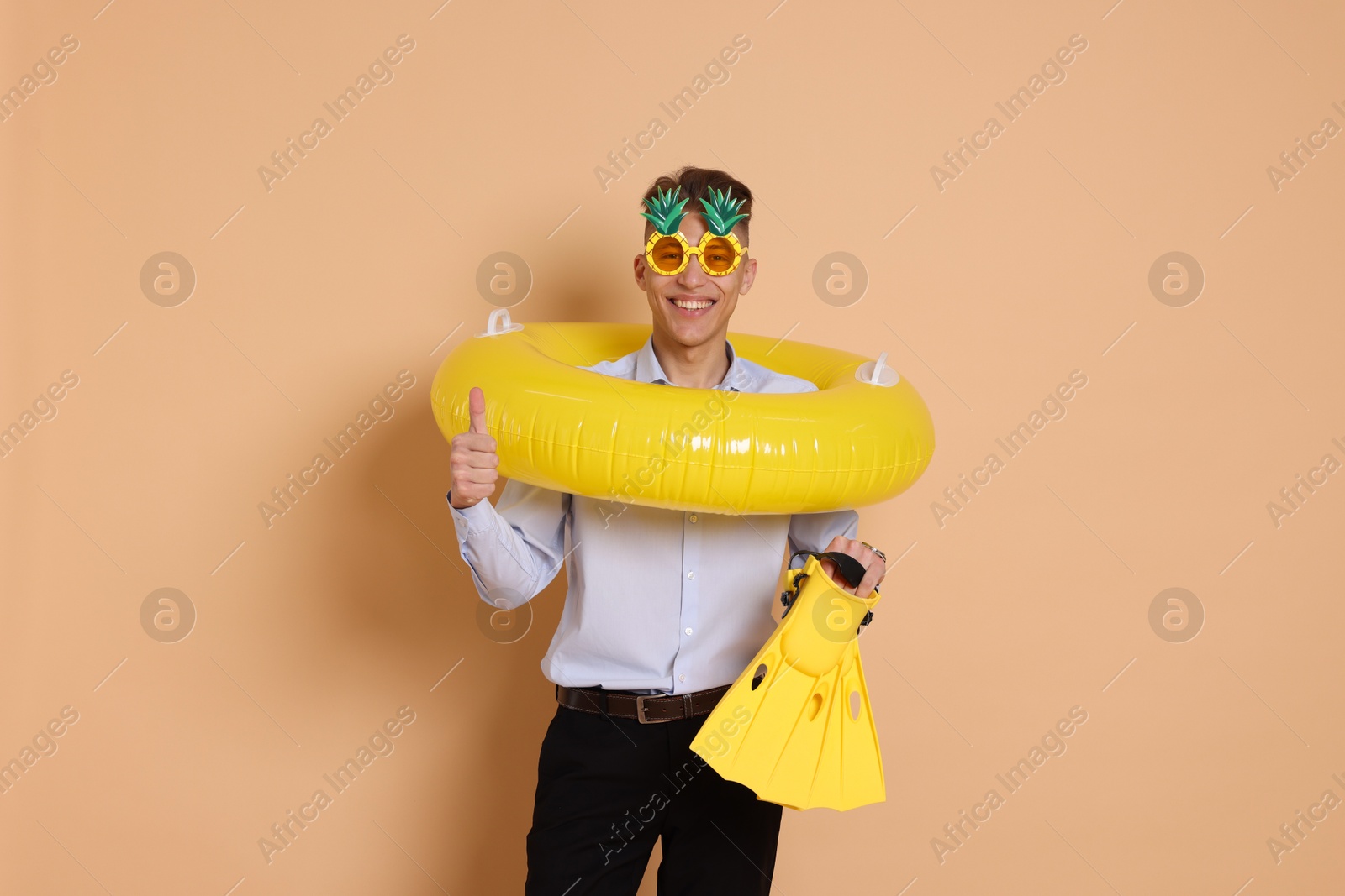 Photo of Businessman with inflatable ring, funny sunglasses and flippers showing thumbs up on beige background