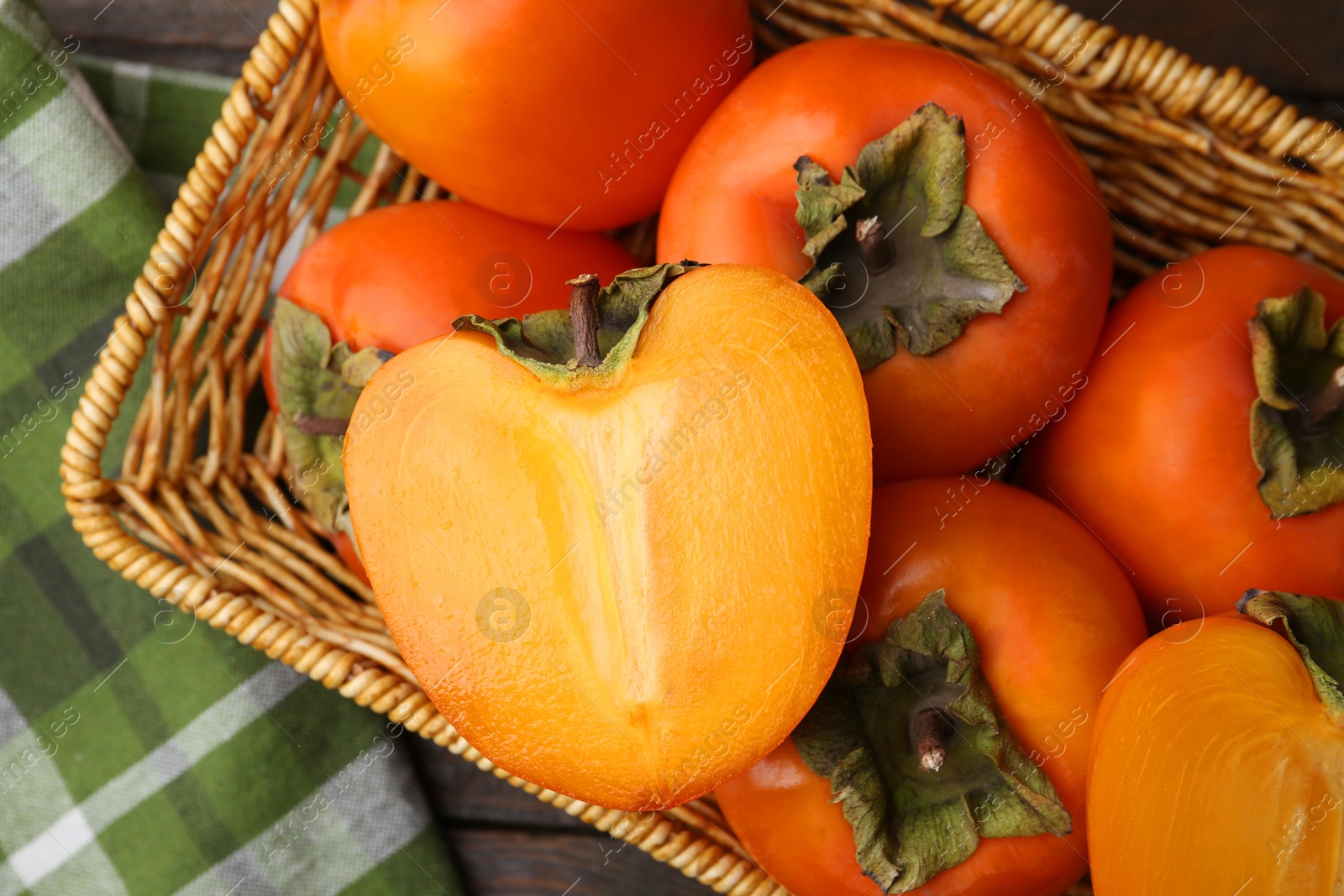 Photo of Whole and cut fresh persimmons in wicker basket on wooden table, top view