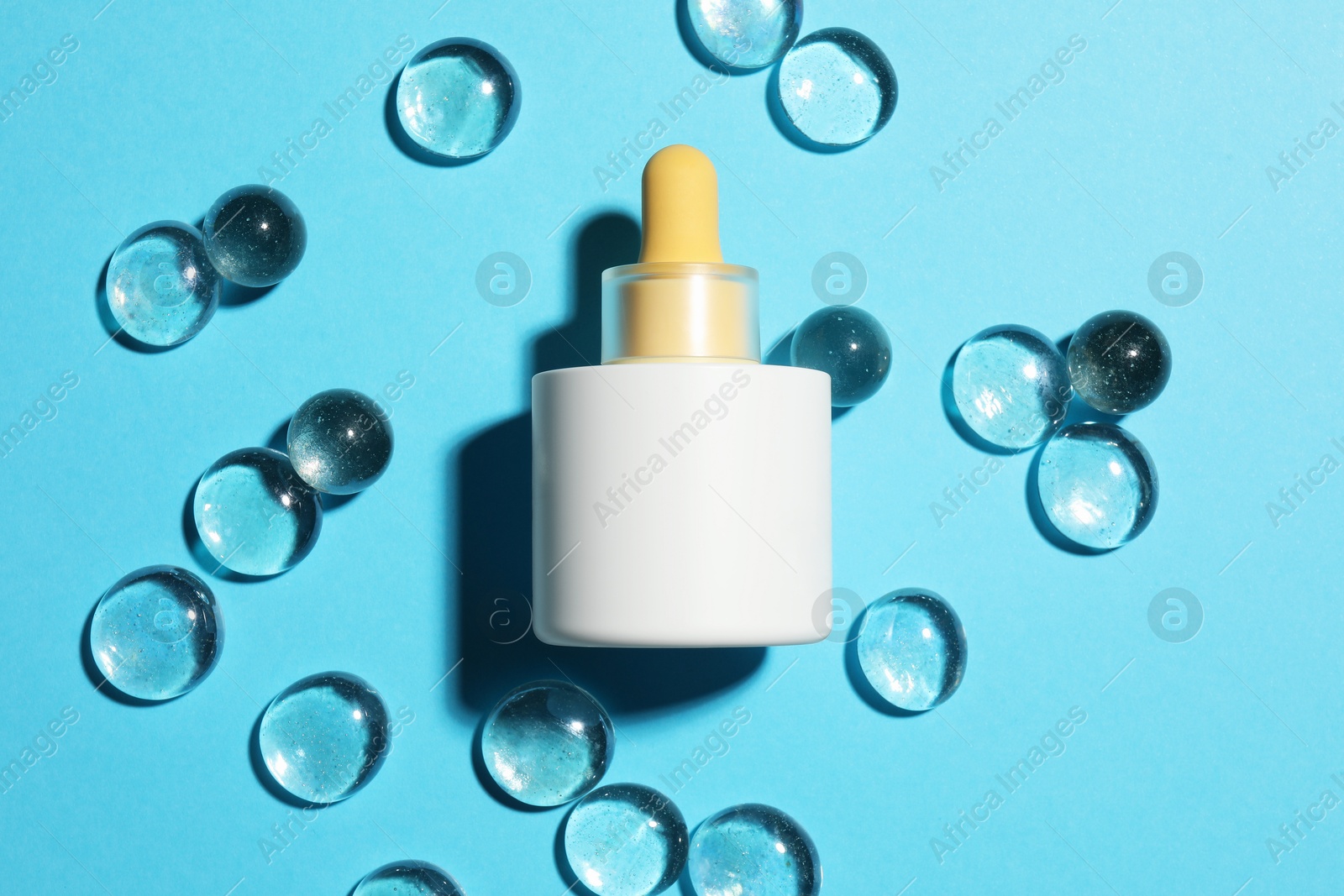 Photo of Bottle of skin care product and transparent decorative stones on light blue background, flat lay