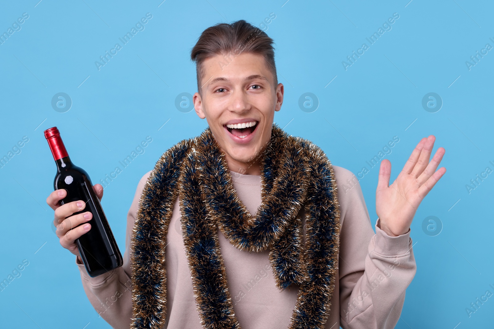 Photo of Happy young man with tinsel and bottle of wine on light blue background