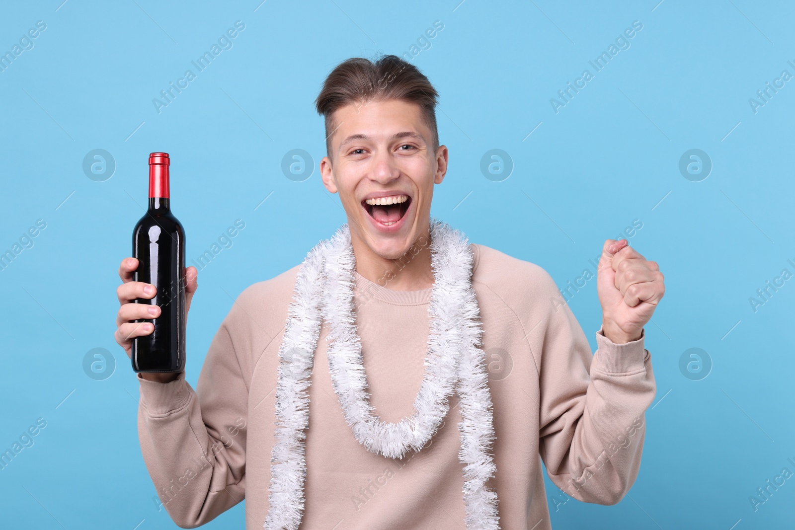 Photo of Happy young man with tinsel and bottle of wine on light blue background