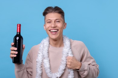 Photo of Happy young man with tinsel and bottle of wine on light blue background