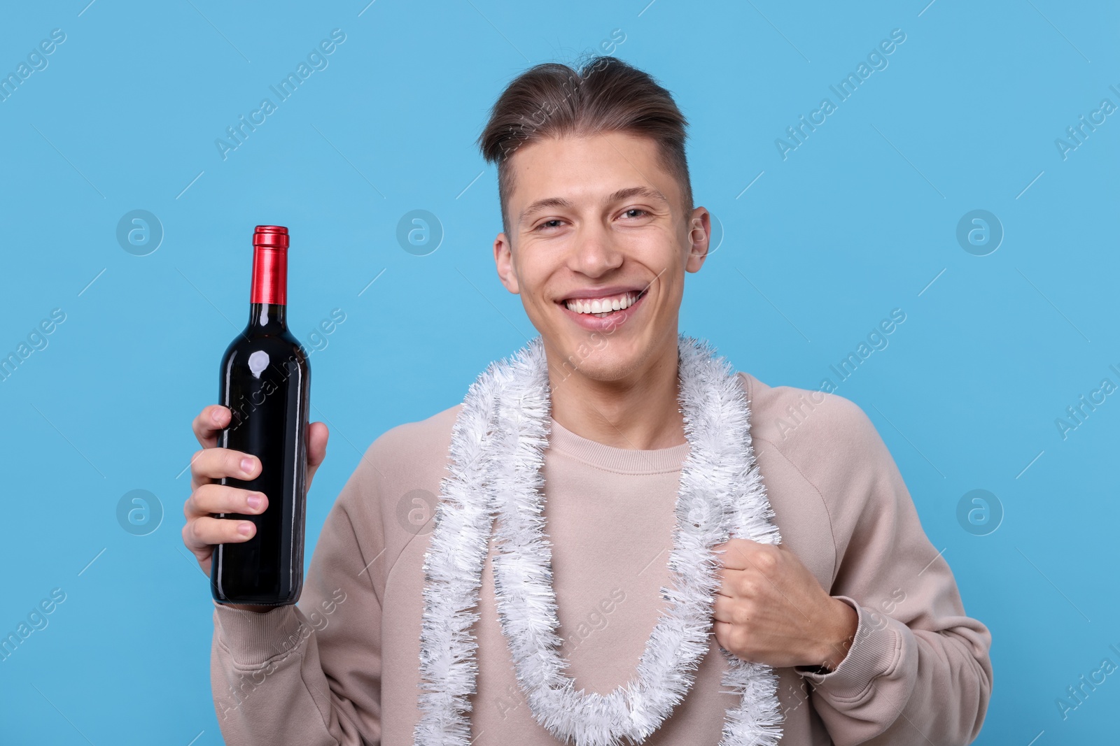 Photo of Happy young man with tinsel and bottle of wine on light blue background