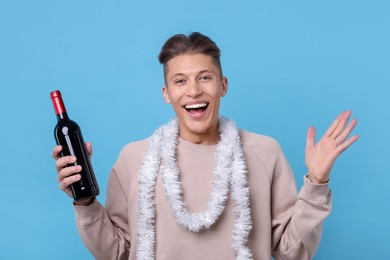 Photo of Happy young man with tinsel and bottle of wine on light blue background