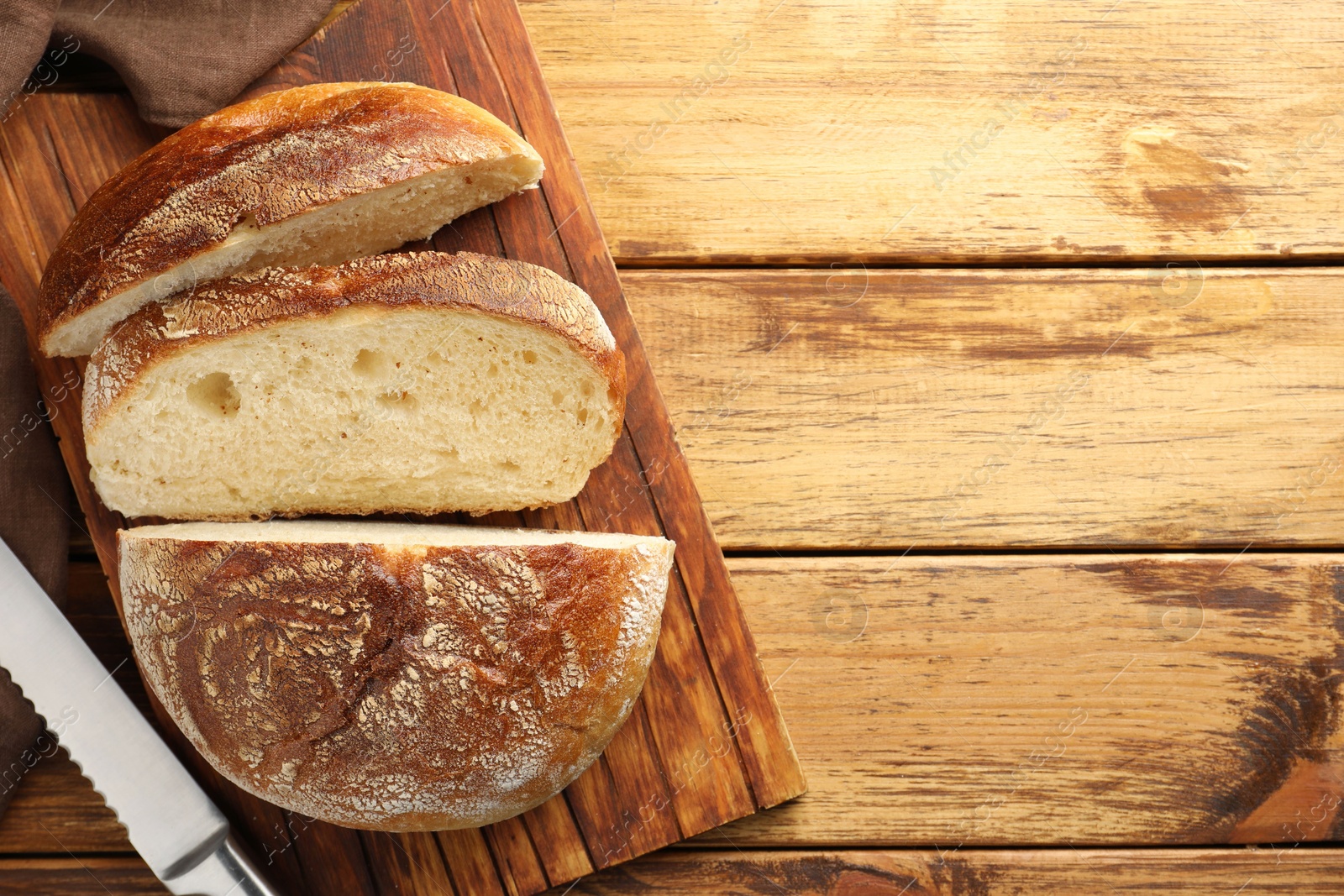 Photo of Cut loaf of fresh bread and knife on wooden table, top view. Space for text