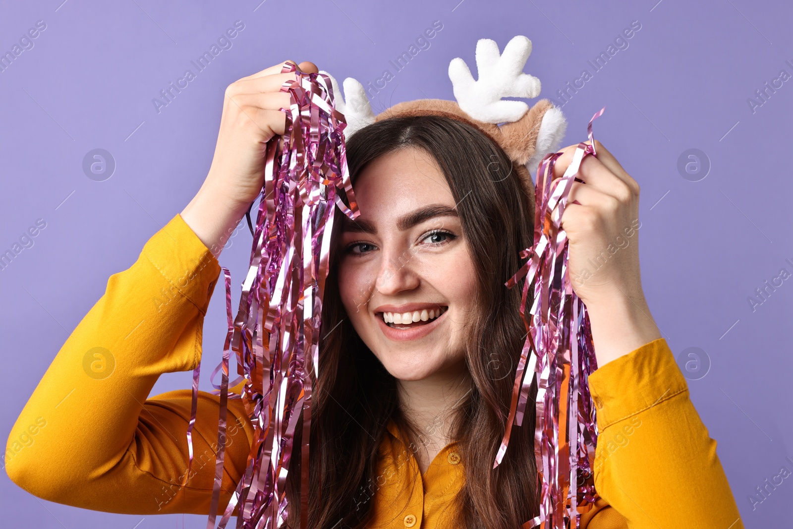 Photo of Happy young woman in reindeer headband with tinsel on purple background