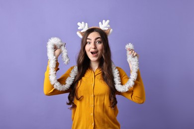 Photo of Emotional young woman in reindeer headband with tinsel on purple background