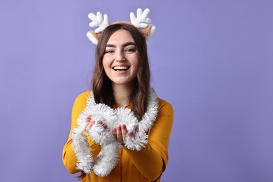 Photo of Happy young woman in reindeer headband with tinsel on purple background. Space for text