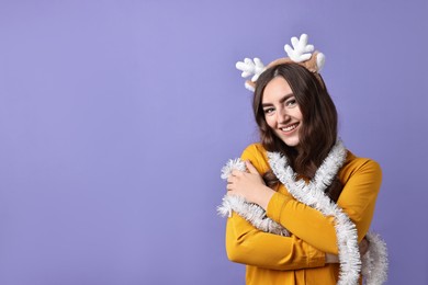 Photo of Happy young woman in reindeer headband with tinsel on purple background. Space for text