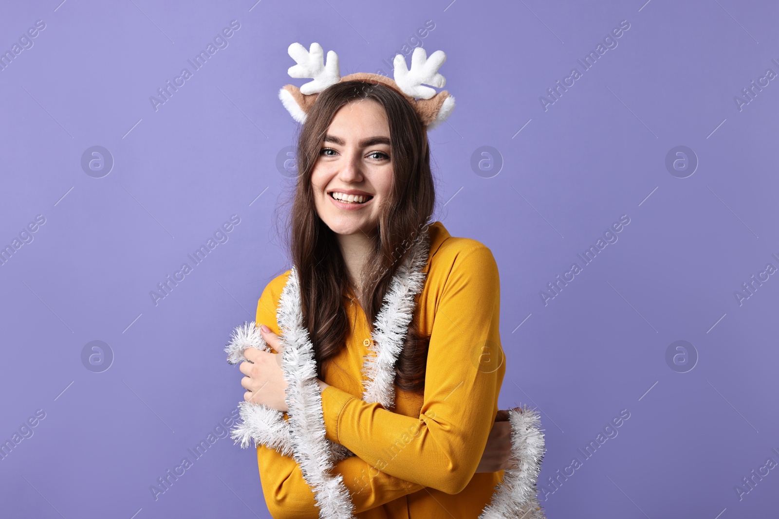 Photo of Happy young woman in reindeer headband with tinsel on purple background