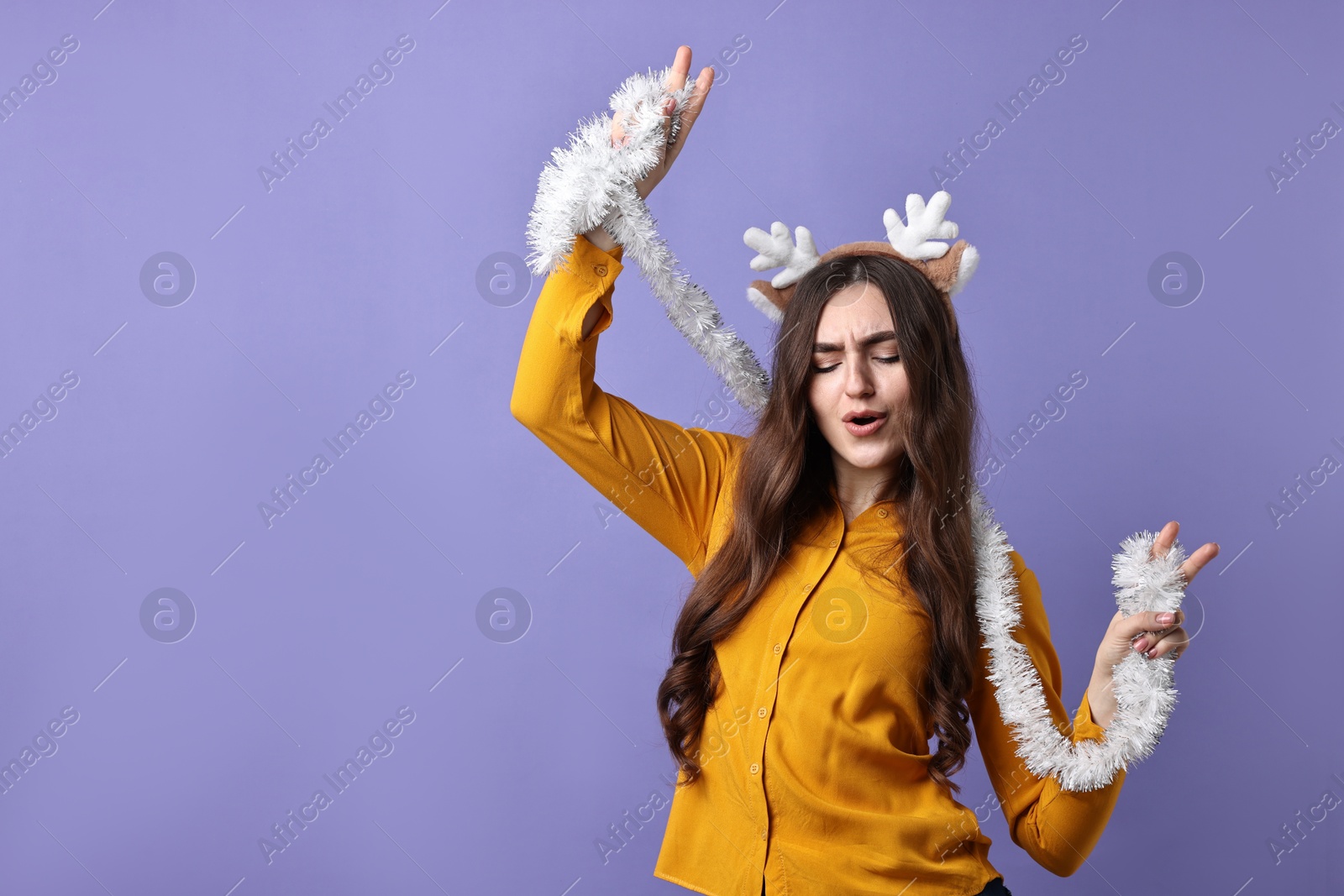 Photo of Emotional young woman in reindeer headband with tinsel on purple background. Space for text