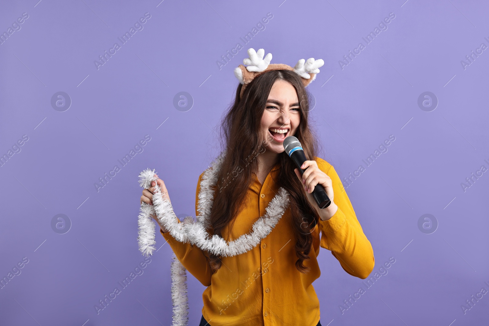 Photo of Young woman in reindeer headband with tinsel singing on purple background