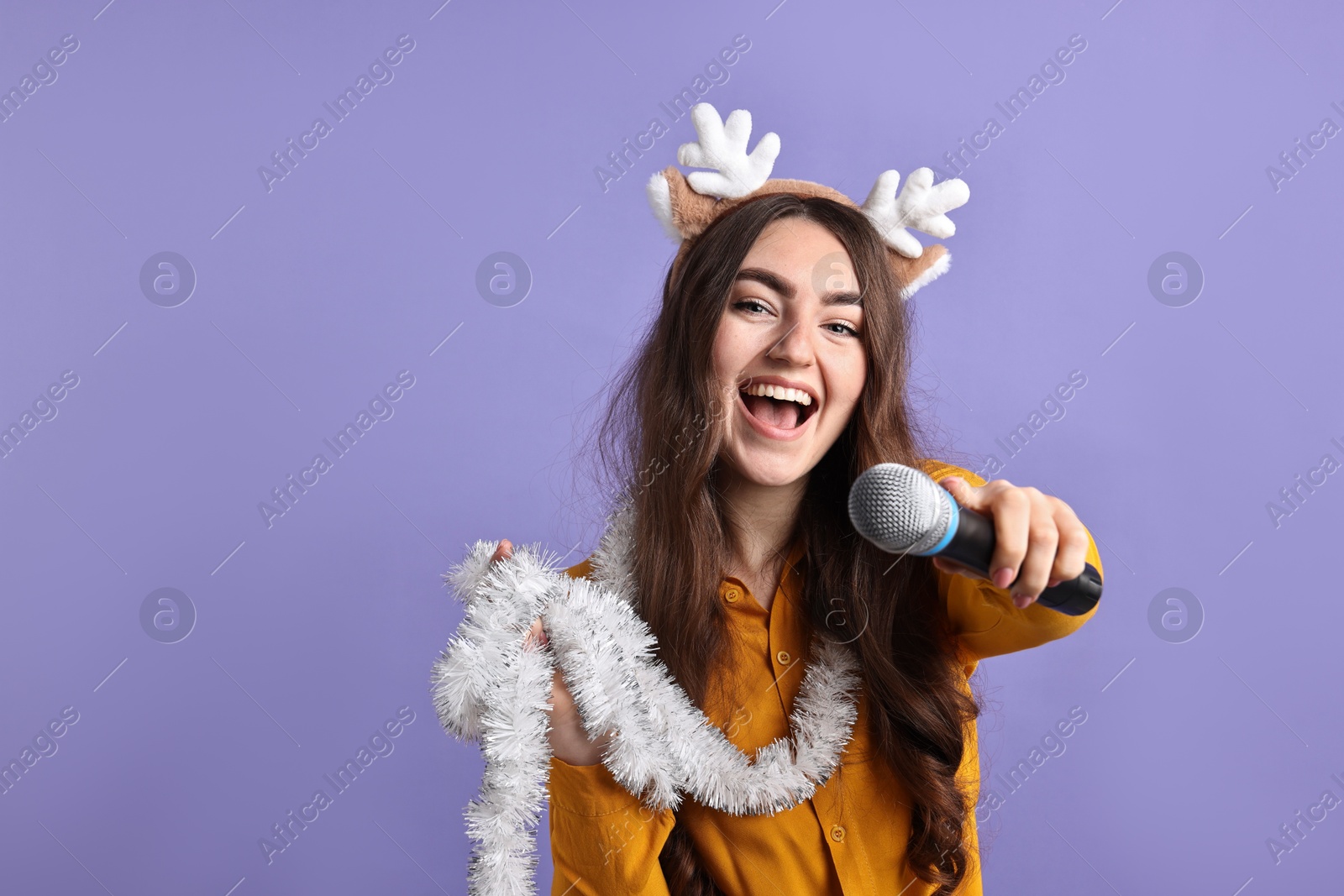 Photo of Happy young woman in reindeer headband with tinsel and microphone on purple background. Space for text