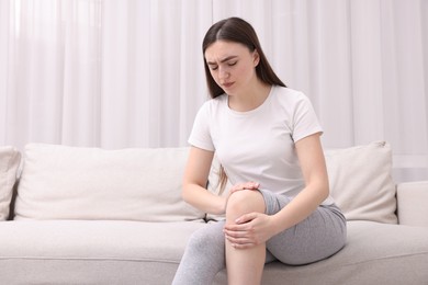 Photo of Young woman suffering from pain in knee on sofa at home. Space for text