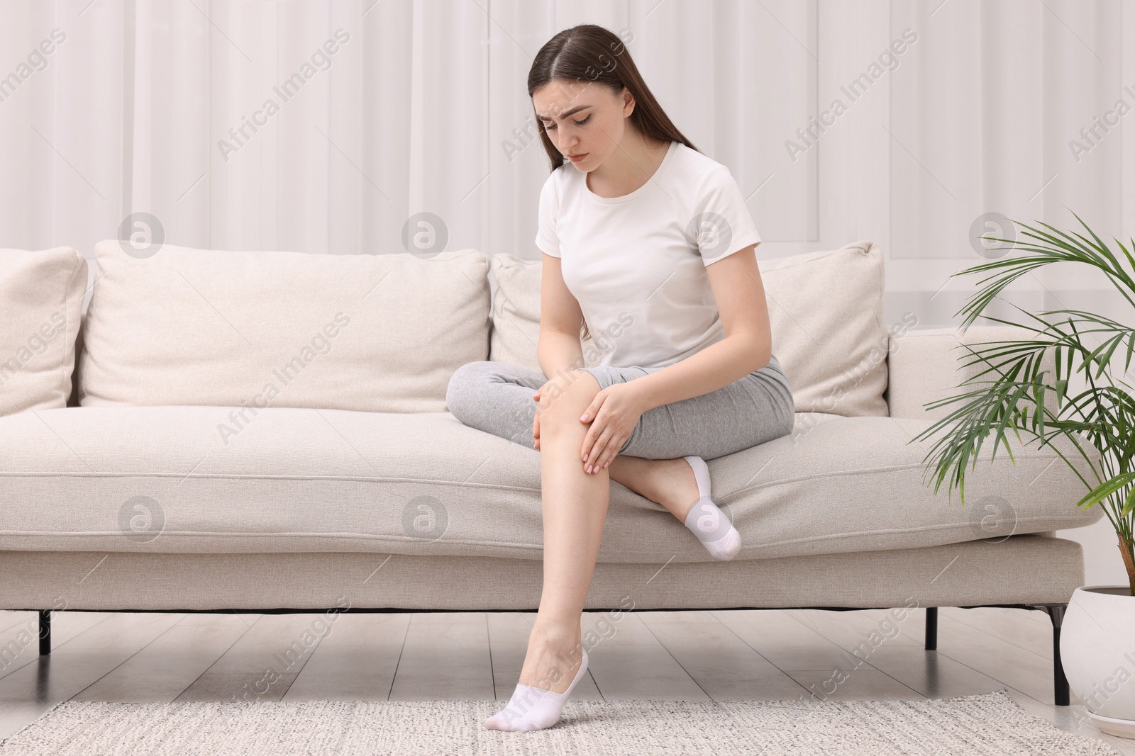 Photo of Young woman suffering from pain in knee on sofa at home