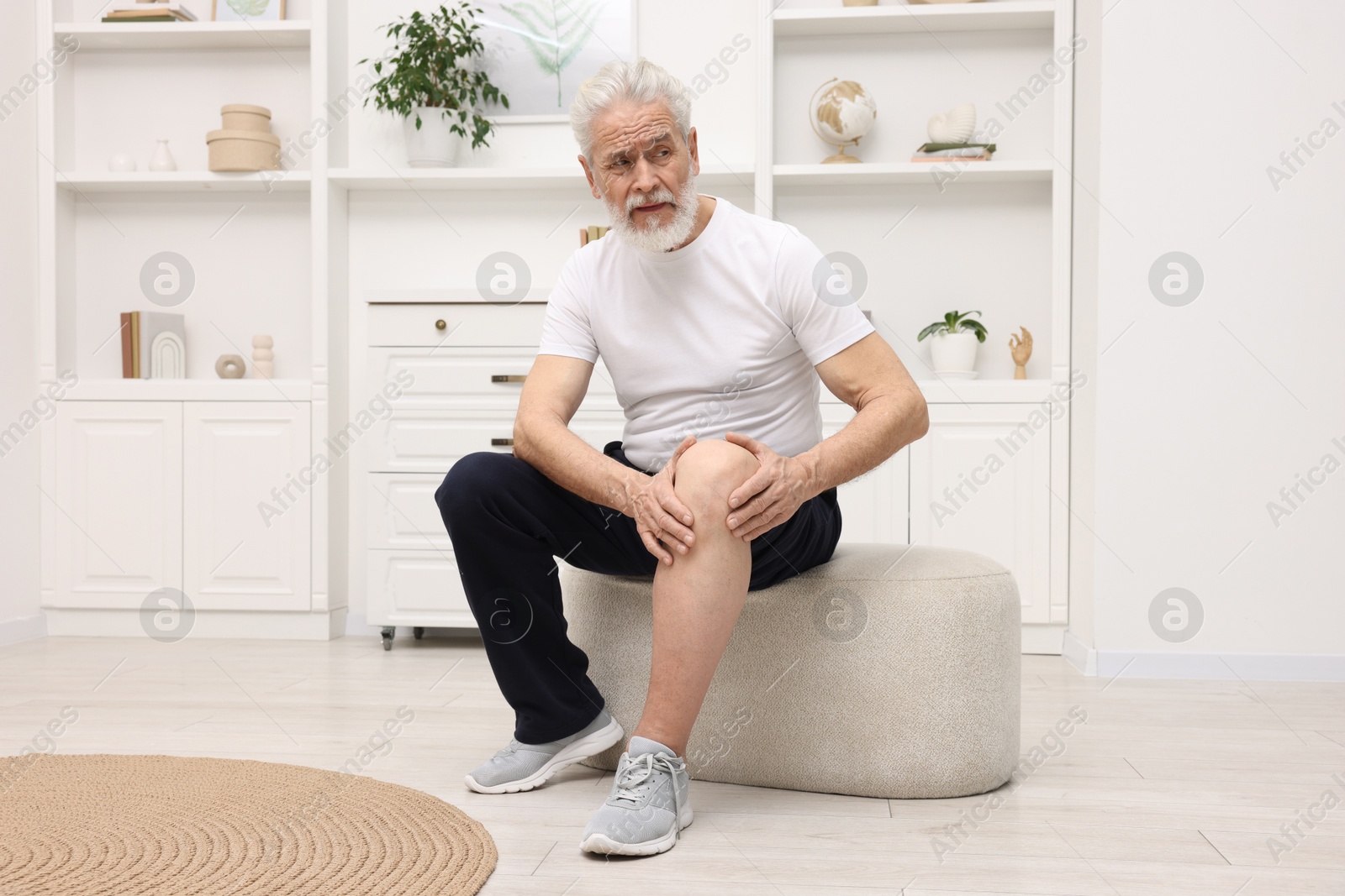 Photo of Senior man suffering from knee pain on pouf at home