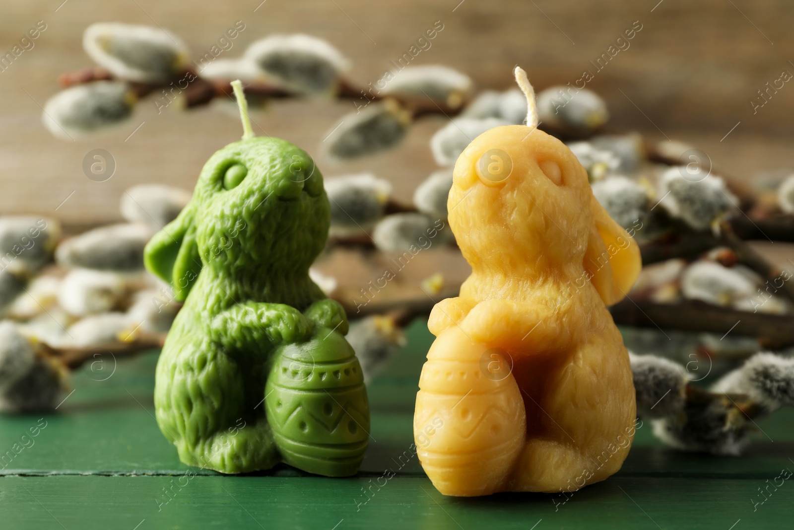 Photo of Colorful bunny-shaped candles and willow branches on green wooden table, closeup. Easter decor