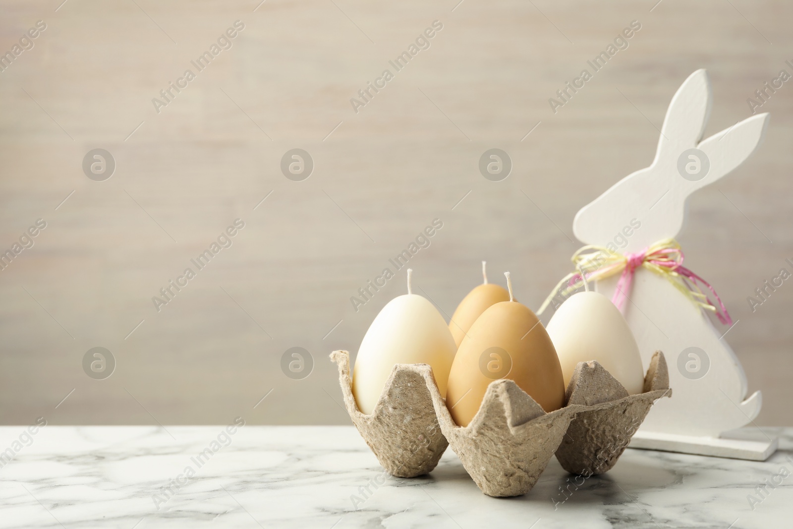 Photo of Egg-shaped candles in carton and bunny figure on white marble table against light gray background, space for text. Easter decor