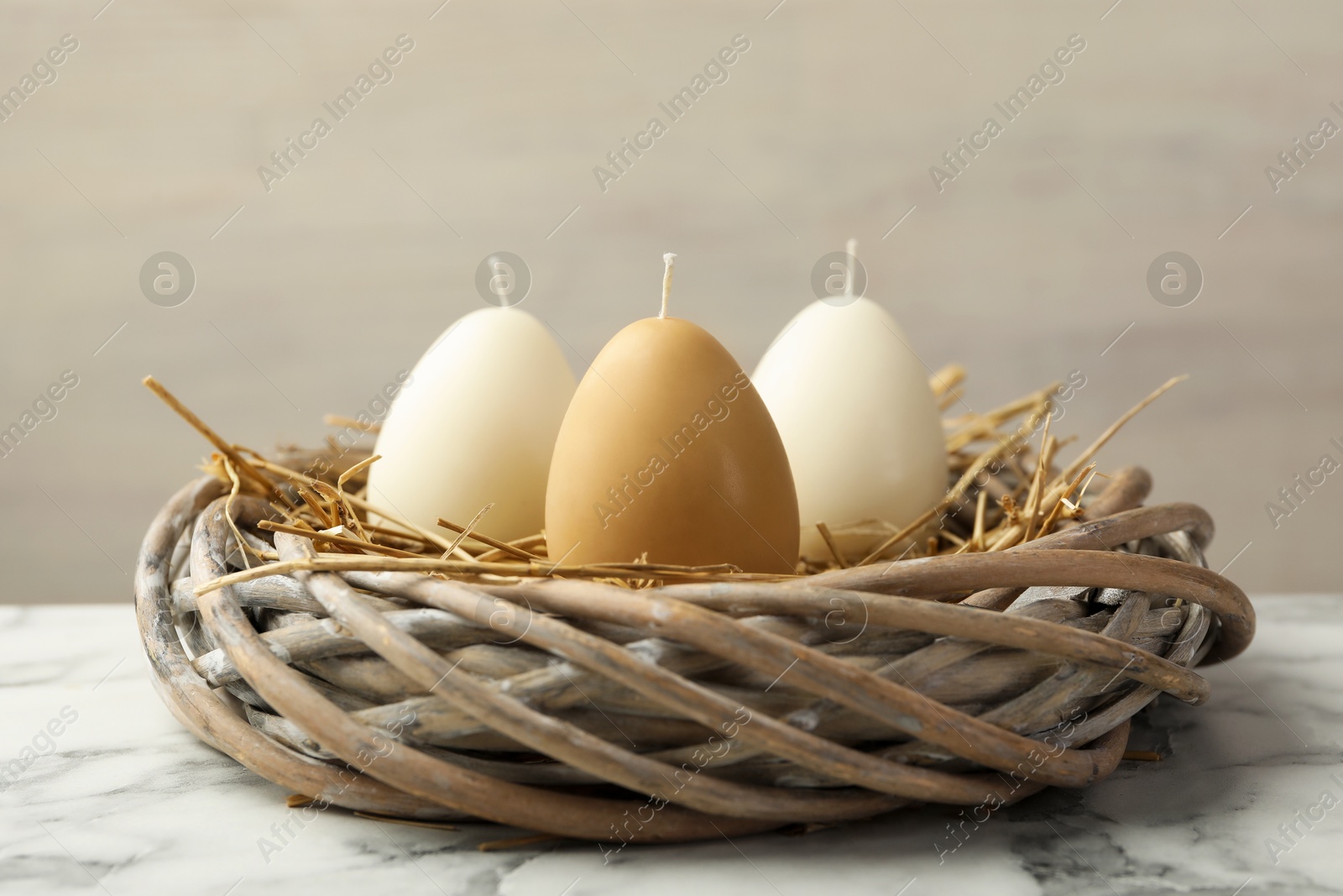 Photo of Egg-shaped candles in nest on white marble table against light gray background, closeup. Easter decor