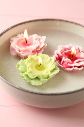 Photo of Beautiful burning flower shaped candles in dish with water on pink wooden table, closeup