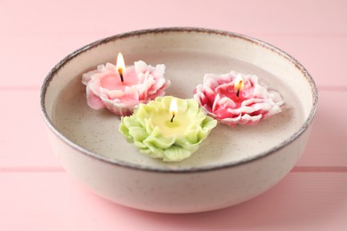 Photo of Beautiful burning flower shaped candles in dish with water on pink wooden table, closeup