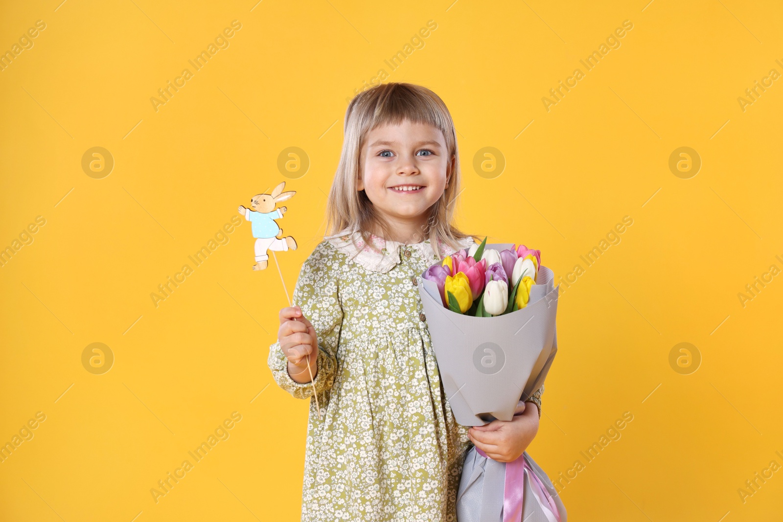 Photo of Smiling little girl with bouquet of tulips and decorative Easter bunny on orange background