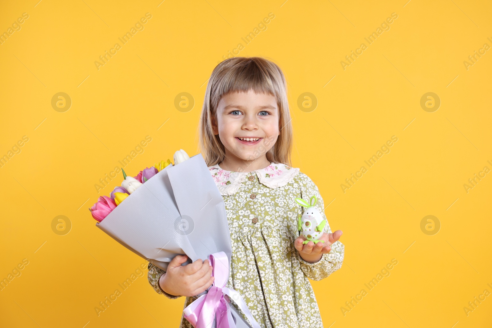 Photo of Smiling little girl with bouquet of tulips and decorative Easter egg on orange background