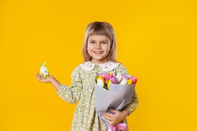 Photo of Smiling little girl with bouquet of tulips and decorative Easter egg on orange background