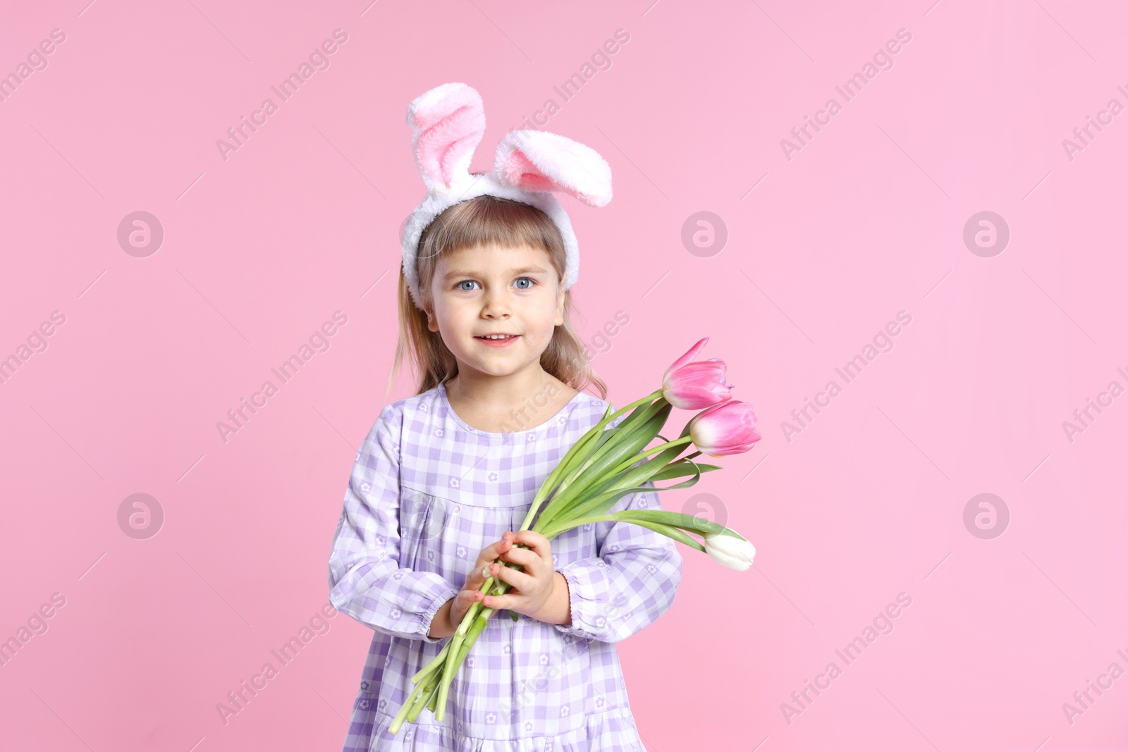 Photo of Smiling little girl in headband with bunny ears holding bouquet of tulips on pink background. Easter celebration