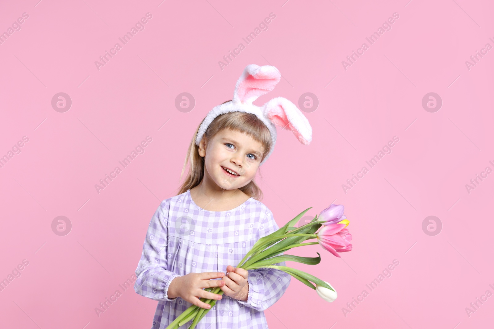 Photo of Smiling little girl in headband with bunny ears holding bouquet of tulips on pink background. Easter celebration