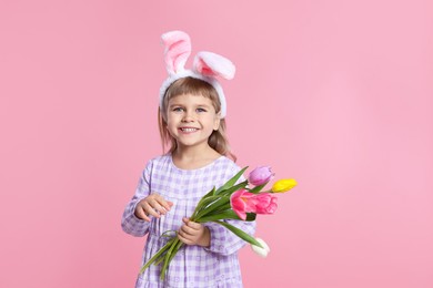 Photo of Smiling little girl in headband with bunny ears holding bouquet of tulips on pink background. Easter celebration