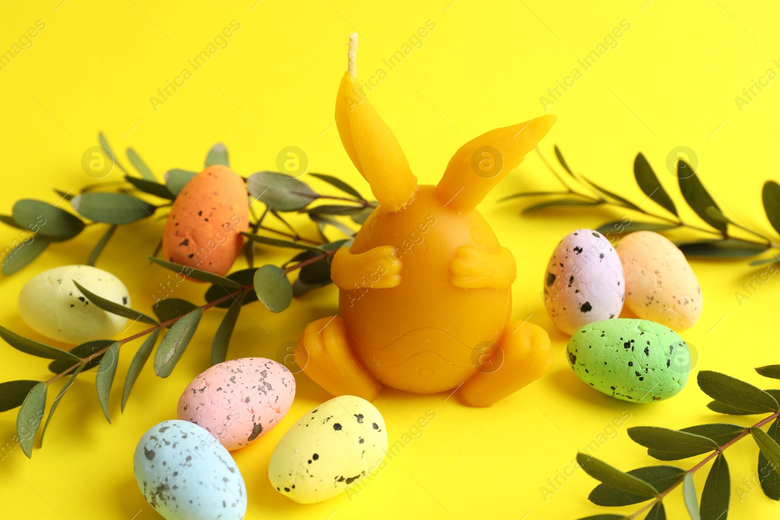 Photo of Beautiful candle, candies and leaves on yellow background, closeup. Easter decor