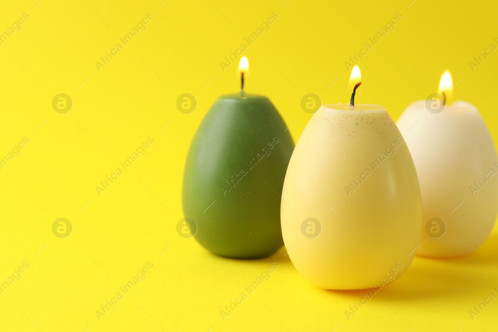 Photo of Beautiful burning egg shaped candles on yellow background, closeup with space for text. Easter decor