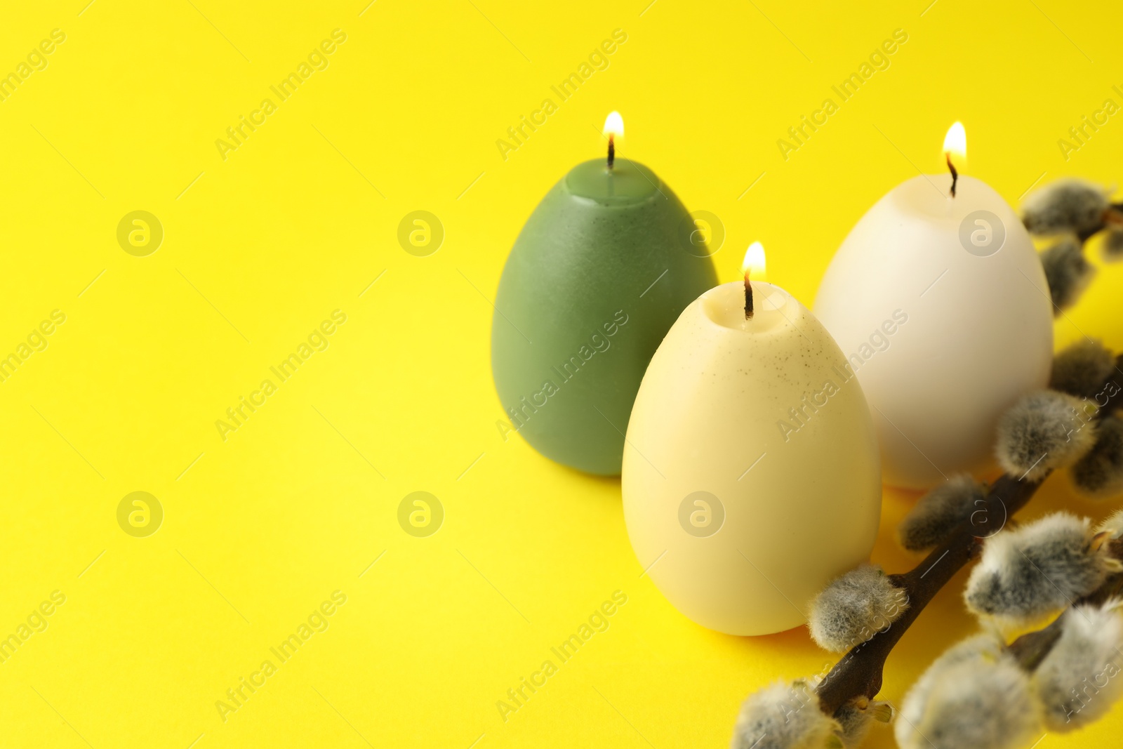 Photo of Beautiful burning egg shaped candles and pussy willow on yellow background, closeup with space for text. Easter decor