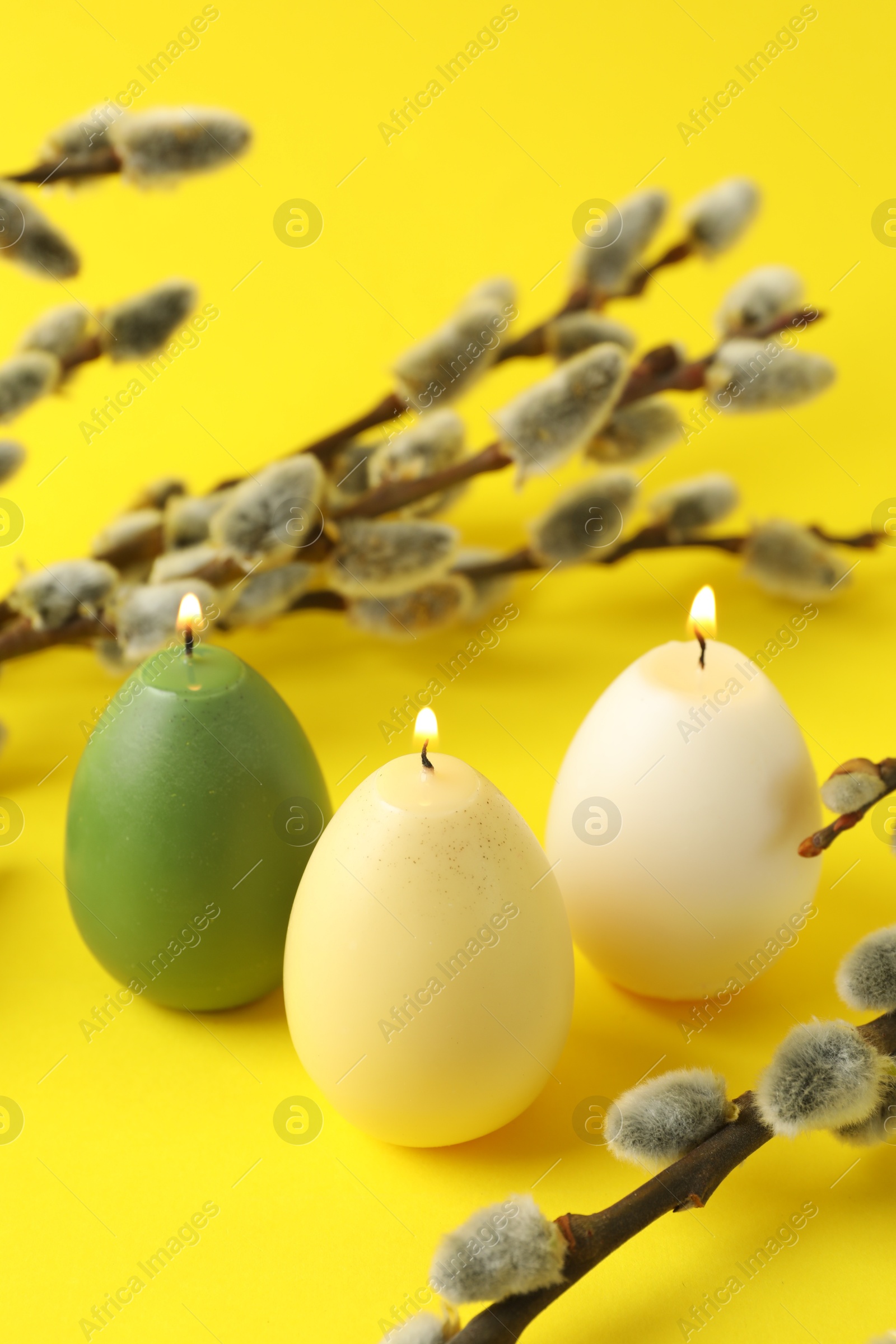 Photo of Beautiful burning egg shaped candles and pussy willow on yellow background, closeup. Easter decor