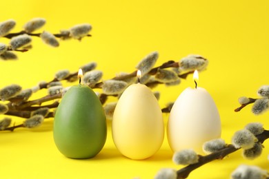 Photo of Beautiful burning egg shaped candles and pussy willow on yellow background, closeup. Easter decor