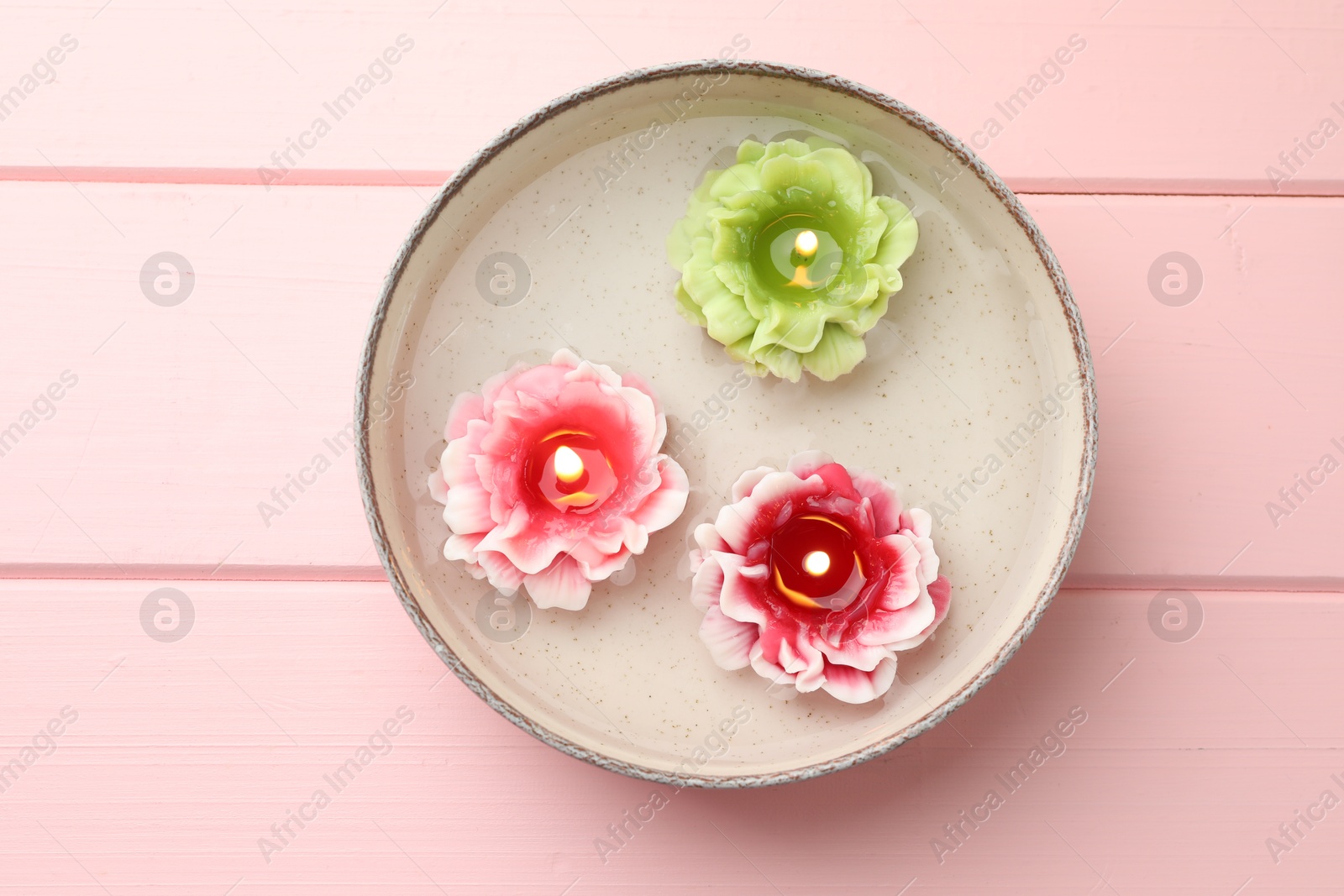 Photo of Beautiful burning flower shaped candles in dish with water on pink wooden table, top view