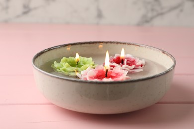 Photo of Beautiful burning flower shaped candles in dish with water on pink wooden table, closeup