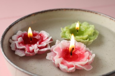 Photo of Beautiful burning flower shaped candles in dish with water on pink table, closeup