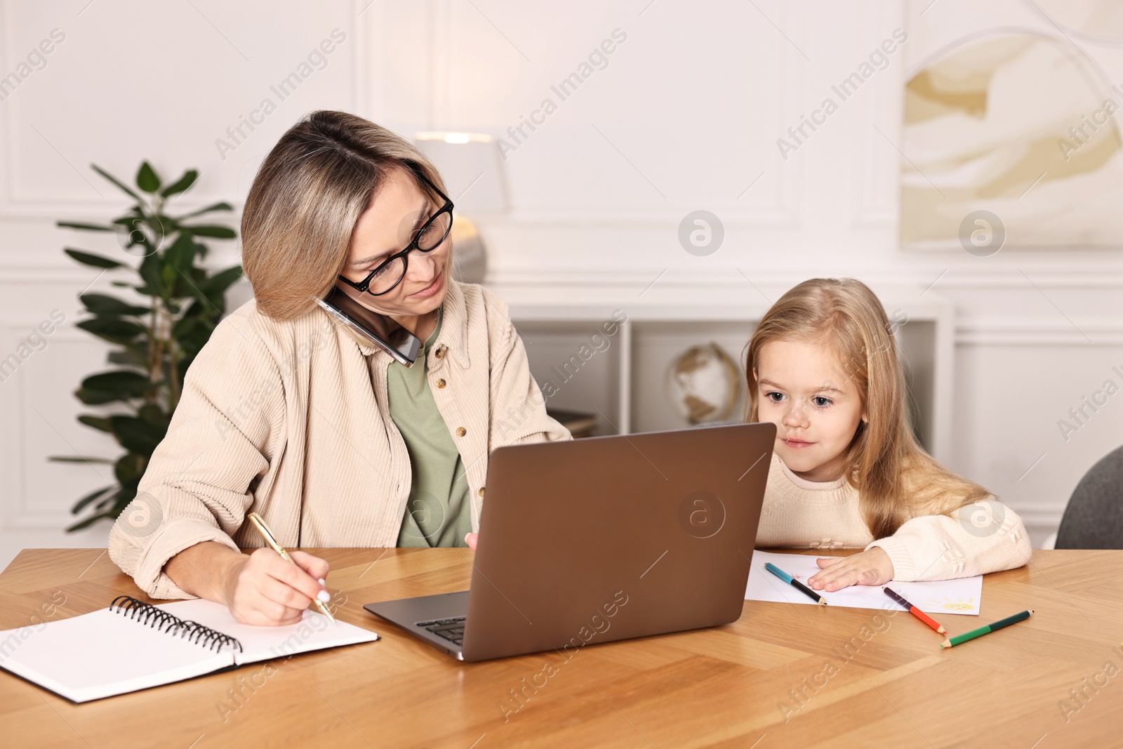 Photo of Work-family balance. Single mother talking by smartphone and her daughter drawing at wooden table indoors