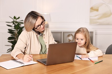 Photo of Work-family balance. Single mother talking by smartphone and her daughter drawing at wooden table indoors