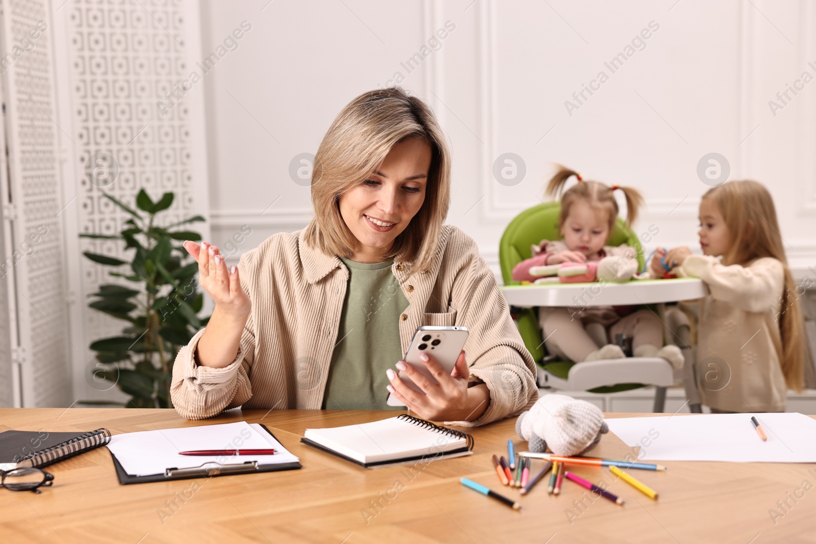 Photo of Work at home. Smiling single mother having videochat by smartphone while her children playing indoors, selective focus